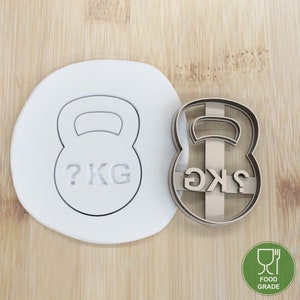 May include: A white plastic cookie cutter in the shape of a kettlebell with the text "? KG" on it. A brown metal cookie cutter in the shape of a kettlebell with the text "? KG" on it. Both cookie cutters are food grade.