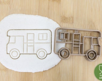 May include: A camper cookie cutter and a camper-shaped cookie on a wooden surface. The cookie cutter is made of metal and has a detailed design of a recreational vehicle. The cookie is imprinted with the same design. The image includes a food grade symbol.