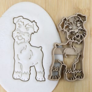 Könnte beinhalten: Ein brauner Plastik-Ausstecher in Form eines Schnauzer-Hundes. Der Ausstecher wird neben einem weißen Stück Modelliermasse gezeigt, in dem die Umrisse des Hundes ausgeschnitten sind.