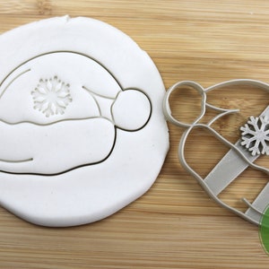 Puede incluir: Un cortador de galletas de plata en forma de gorro de Pap&aacute; Noel con un copo de nieve en la parte delantera. El cortador est&aacute; sobre una superficie de madera. Grado alimenticio.