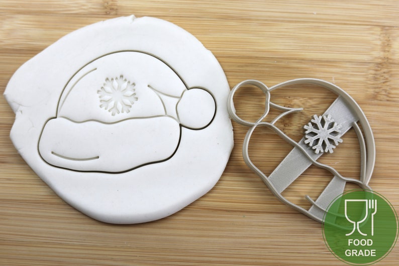 Puede incluir: Un cortador de galletas de plata en forma de gorro de Pap&aacute; Noel con un copo de nieve en la parte delantera. El cortador est&aacute; sobre una superficie de madera. Grado alimenticio.