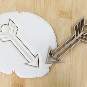 May include: A brown plastic cookie cutter in the shape of an arrow with a geometric pattern. The cutter is on a wooden surface and has been used to cut out a shape in a piece of white dough.