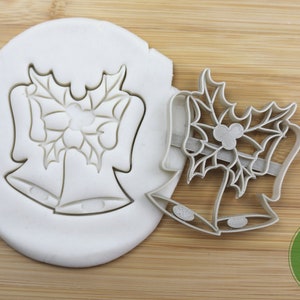 Puede incluir: Un cortador de galletas de metal con forma de dos campanas con hojas de acebo y bayas. El cortador es de calidad alimentaria y se muestra sobre una superficie de madera.