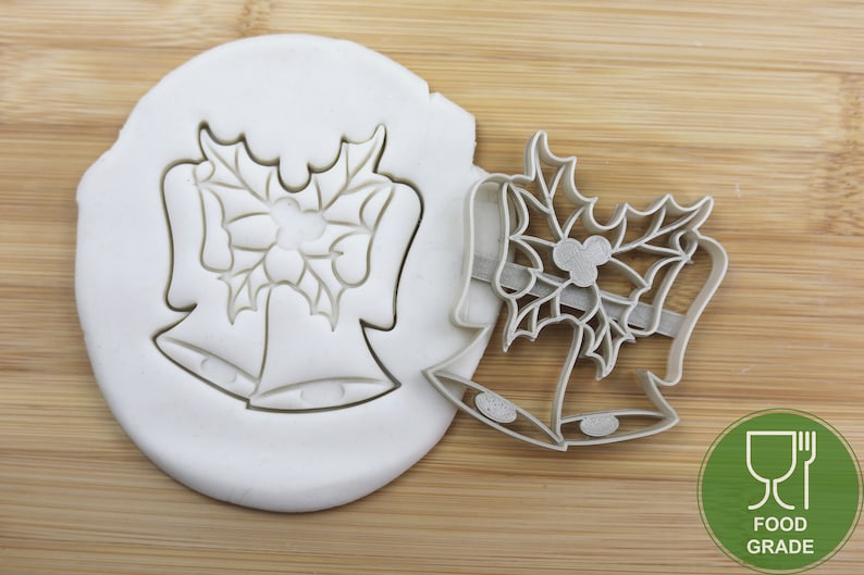 Puede incluir: Un cortador de galletas de metal con forma de dos campanas con hojas de acebo y bayas. El cortador es de calidad alimentaria y se muestra sobre una superficie de madera.