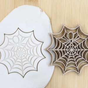 Könnte beinhalten: Ein brauner Plastik-Ausstecher in Form eines Spinnennetzes mit einer Spinne in der Mitte. Der Ausstecher ist lebensmittelecht und wird auf einer weißen Oberfläche gezeigt.