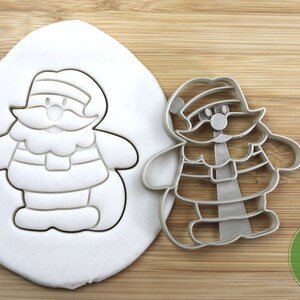 Puede incluir: Un cortador de galletas de plata con forma de Pap&aacute; Noel de dibujos animados. El cortador est&aacute; sobre una superficie de madera con una impresi&oacute;n blanca del cortador en un bloque de plastilina. El cortador est&aacute; etiquetado como "Calidad alimentaria".