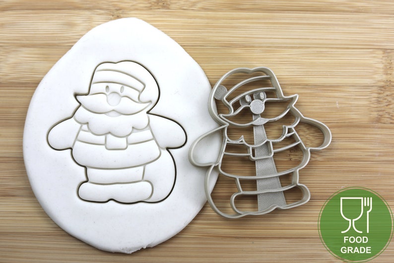 Puede incluir: Un cortador de galletas de plata con forma de Pap&aacute; Noel de dibujos animados. El cortador est&aacute; sobre una superficie de madera con una impresi&oacute;n blanca del cortador en un bloque de plastilina. El cortador est&aacute; etiquetado como "Calidad alimentaria".