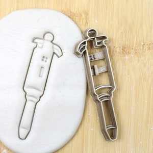 Op de afbeelding: Een zilveren koekjesvormpje in de vorm van een pipet, een laboratoriumgereedschap dat wordt gebruikt om kleine hoeveelheden vloeistof over te brengen. De vormpje is te zien op een houten oppervlak naast een stuk witte modelleerklei dat met de vormpje is geperst.