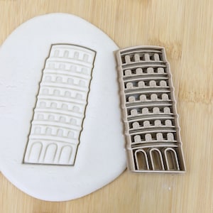 May include: A brown plastic cookie cutter shaped like the Leaning Tower of Pisa. The cutter is on a wooden surface next to a white piece of modeling clay with the cutter impression in it. The cutter is labeled "Food Grade".