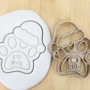 Könnte beinhalten: Ein brauner Plastik-Ausstecher in Form einer Hundepfote, die einen Weihnachtsmann-Hut trägt. Die Pfote hat die Worte "I love dog" im Inneren. Der Ausstecher befindet sich auf einer Holzoberfläche.