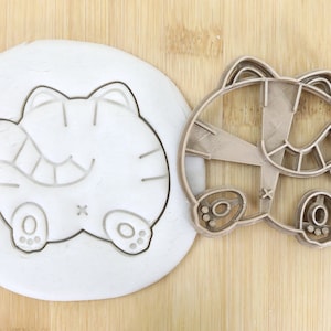 May include: A brown plastic cookie cutter in the shape of a cartoon cat. The cutter is on a wooden surface next to a white piece of dough with the outline of the cat cut into it. The cutter is labeled "Food Grade".