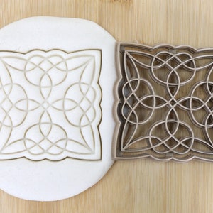 Könnte beinhalten: Ein brauner, lebensmittelechter Plastik-Ausstecher in Form eines Quadrats mit einem keltischen Knotenmuster. Der Ausstecher wird auf einer Holzoberfläche gezeigt, mit einem Muster, das in einen weißen Teig gedrückt wurde.