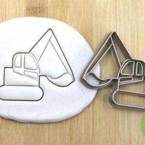 Könnte beinhalten: Ein silberner Keksausstecher in Form eines Baggers. Der Ausstecher wird neben einem weißen Fondant-Abdruck des Baggers gezeigt.