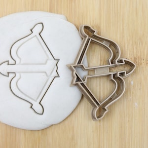 Könnte beinhalten: Ein goldfarbener Metall-Ausstecher in Form von Pfeil und Bogen. Der Ausstecher ist auf einer Holzoberfläche neben einem Stück weißen Modelliertones mit dem Abdruck des Ausstechers zu sehen.