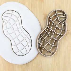 Puede incluir: Un cortador de galletas de plástico marrón en forma de cacahuete con un diseño detallado. El cortador se muestra sobre una superficie de madera junto a una pieza de plastilina blanca con la impresión del cortador.