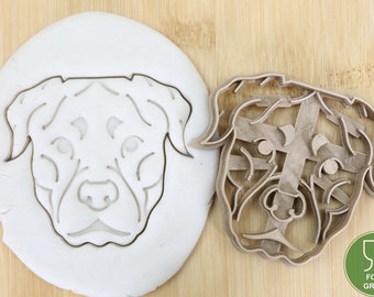 Könnte beinhalten: Ein brauner Plastik-Ausstecher in Form eines HundeKopfes. Der Ausstecher liegt auf einer Holzoberfläche neben einem weißen Keksausstecher aus Teig in Form desselben Hundes.