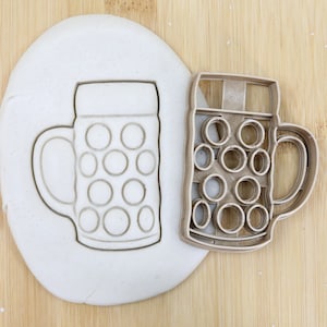 Könnte beinhalten: Nahaufnahme eines braunen Plastik-Ausstechers in Form eines Bierkruges mit Henkel. Der Ausstecher liegt auf einer Holzoberfläche neben einem Stück weißen Modelliertones, aus dem die gleiche Form ausgeschnitten ist. Der Ausstecher ist mit "Food Grade" gekennzeichnet.