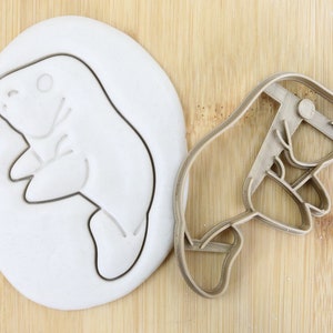 Peut inclure: Un emporte-pièce en forme de lamantin à côté d'un biscuit en forme de lamantin dans une pâte blanche. L'emporte-pièce est en matériau marron clair. L'image est sur une surface en bois brun clair. Un symbole vert de qualité alimentaire est en bas à droite.