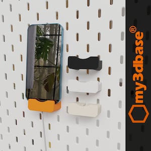 Könnte beinhalten: Eine weiße Lochwand mit drei Telefonhaltern. Die Telefonhalter sind schwarz, weiß und orange. Die Telefonhalter sind so konzipiert, dass sie ein Telefon vertikal halten. Die Lochwand hat ein Logo mit der Aufschrift "my3dbase" in orange.