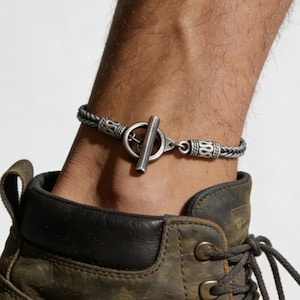 May include: A silver ankle bracelet with a toggle clasp is worn above a brown leather boot. The bracelet has a braided design and is secured with a T-bar clasp. The person is wearing blue jeans.