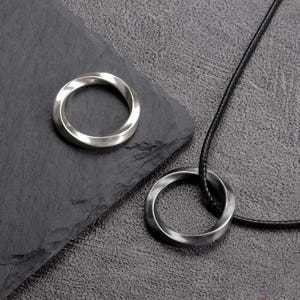 May include: A silver-colored ring and a matching pendant necklace with a twisted design. The pendant hangs from a black cord. The ring sits on a dark gray surface.