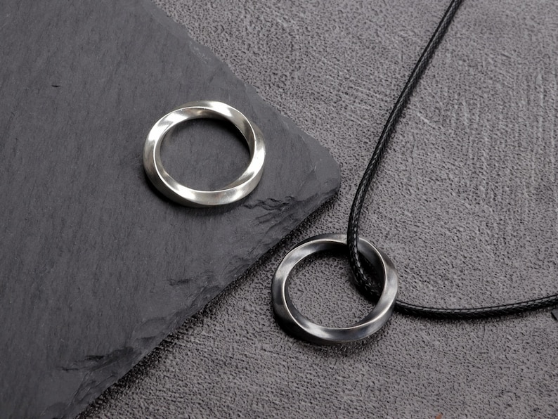 May include: A silver-colored ring and a matching pendant necklace with a twisted design. The pendant hangs from a black cord. The ring sits on a dark gray surface.
