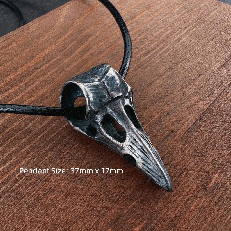 May include: A silver-colored raven skull pendant on a black cord. The pendant is 37mm x 17mm. The skull has detailed features and is set against a wooden background. The pendant is a gothic or alternative style.