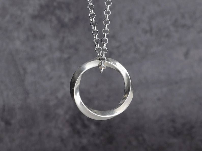 May include: A silver-colored pendant necklace featuring a Mobius strip design. The pendant is a continuous loop, suspended from a delicate chain. The necklace is displayed against a gray background, highlighting the polished metal.