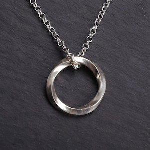 May include: A silver-toned necklace with a twisted circular pendant. The pendant is suspended from a delicate chain. The jewelry is displayed against a dark, textured background.