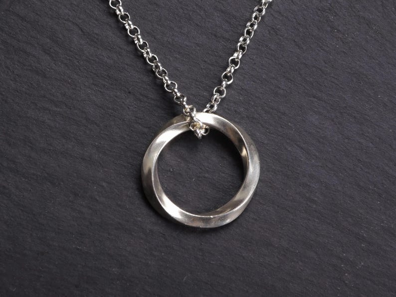 May include: A silver-toned necklace with a twisted circular pendant. The pendant is suspended from a delicate chain. The jewelry is displayed against a dark, textured background.