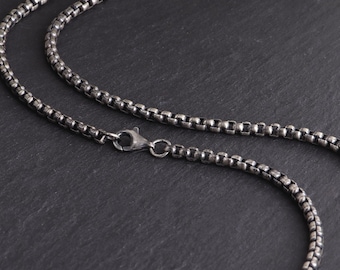 May include: Close-up of a silver-toned necklace with a box chain design. The necklace features a lobster clasp closure. The chain is made of small, square links, creating a textured appearance. The jewelry is displayed against a dark background.