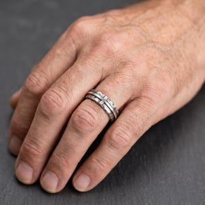Peut inclure: Une bague en argent avec un motif de croix. La bague a une surface texturée et une bande avec une croix au centre. La bague est sur le doigt d'une personne.