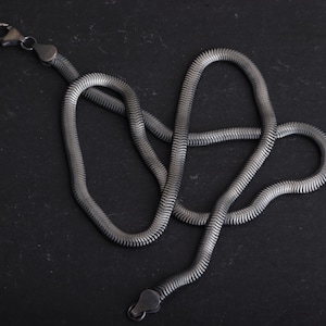 May include: A silver-coloured snake chain necklace with a textured, ribbed design. The necklace is displayed on a dark, textured surface, highlighting its intricate details and the clasp. The chain is a classic jewellery piece.