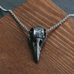 May include: A silver-colored raven skull pendant necklace. The skull pendant is detailed with eye sockets, a beak, and textured surfaces. The necklace chain is a delicate, linked design. The pendant is displayed on a wooden surface.