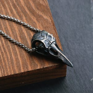 May include: A silver-colored raven skull pendant necklace. The pendant is detailed with textured features and hangs from a delicate chain. The necklace is displayed on a wooden surface, with a dark background.
