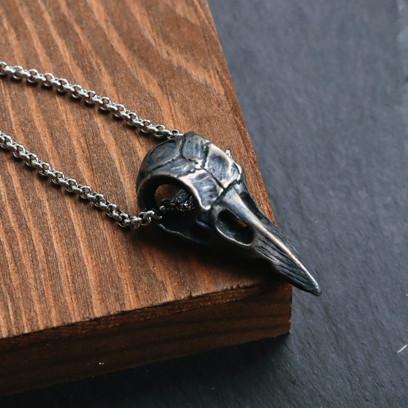 May include: A silver-colored raven skull pendant necklace. The pendant is detailed with textured features and hangs from a delicate chain. The necklace is displayed on a wooden surface, with a dark background.