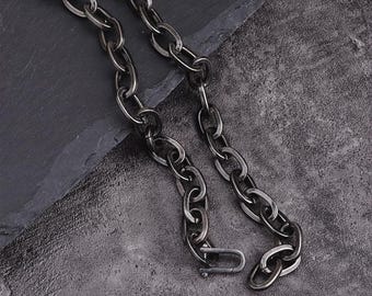 Può includere: Una collana a catena color argento con grandi maglie ovali e una chiusura. La collana è esposta su una superficie strutturata grigio scuro, creando un contrasto con la lucentezza metallica della catena.