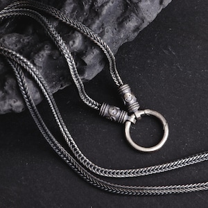 May include: A silver-toned necklace with a braided chain design. The necklace features a circular clasp and decorative cylindrical beads with etched patterns. The chain is displayed on a dark, textured surface.