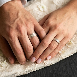 May include: Two silver wedding bands with a Greek key design are displayed on a lace surface. The rings are on the ring fingers of two hands. The hands are resting on a white lace fabric.