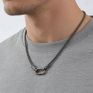 May include: A silver-toned necklace with a woven chain and a rectangular clasp. The necklace is worn against a gray t-shirt, showcasing the jewelry's design and texture. The clasp is a prominent feature.