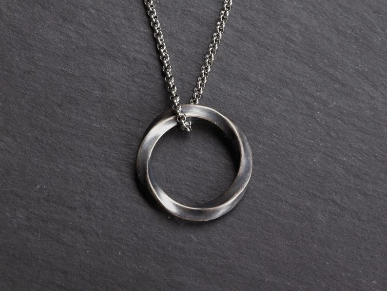 May include: A silver-toned pendant necklace featuring a twisted Mobius strip design. The pendant hangs from a delicate chain. The jewelry is displayed against a dark, textured background.