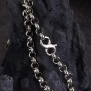Puede incluir: Primer plano de un collar de cadena plateada con cierre de mosquetón. La cadena está hecha de eslabones circulares entrelazados. El collar se muestra sobre un fondo oscuro y texturizado.