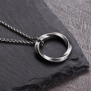 May include: A silver-toned pendant necklace featuring a twisted Mobius strip design. The pendant is suspended from a delicate chain. The jewelry is displayed on a dark, textured surface.