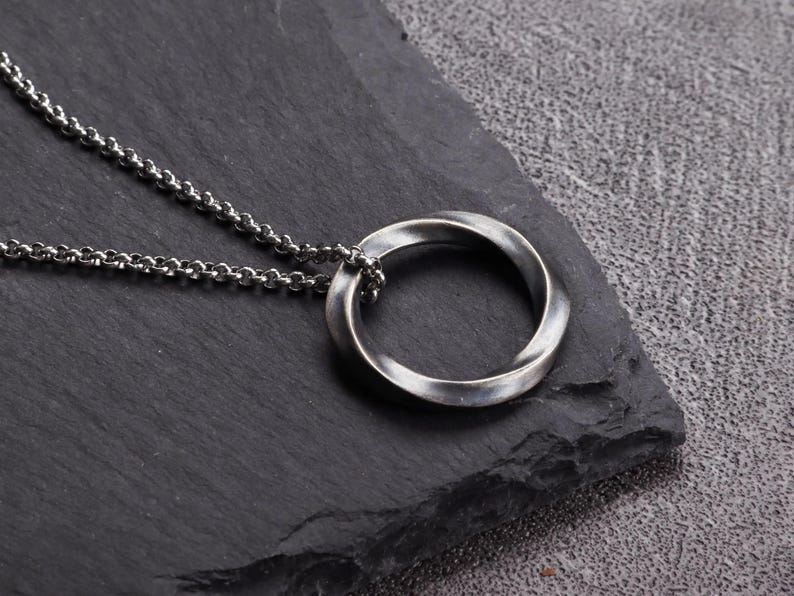 May include: A silver-toned pendant necklace featuring a twisted Mobius strip design. The pendant is suspended from a delicate chain. The jewelry is displayed on a dark, textured surface.