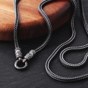 Può includere: Una collana color argento con un design a catena intrecciata. La collana presenta chiusure decorative con dettagli incisi e una chiusura circolare. La catena è esposta su uno sfondo scuro.