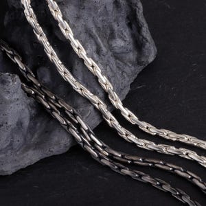 Puede incluir: Primer plano de tres collares de cadena plateados con un diseño texturizado y enlazado. Las cadenas están colocadas sobre una piedra oscura y tosca, sobre un fondo negro. Las cadenas varían ligeramente de color, una de ellas aparece más oscura.