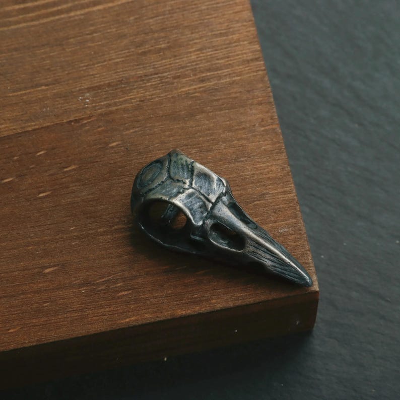 May include: A detailed, silver-toned raven skull pendant. The pendant is intricately designed with visible textures and details, showcasing the shape of the skull and beak. The pendant is placed on a wooden surface.