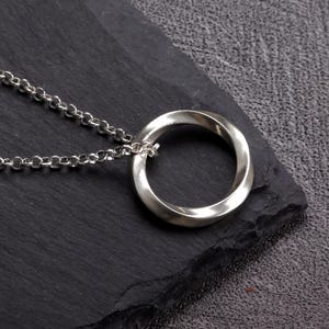 May include: A silver-colored pendant necklace featuring a Mobius strip design. The pendant is a continuous loop, and it hangs from a delicate chain. The necklace is displayed on a dark, textured surface.