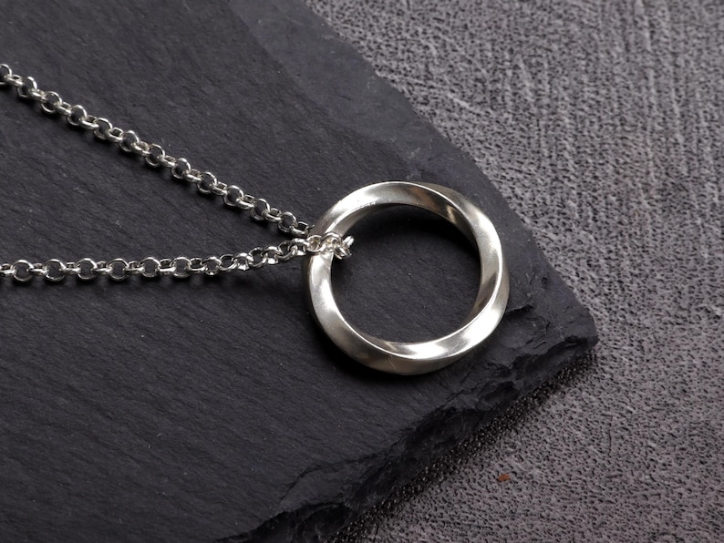 May include: A silver-colored pendant necklace featuring a Mobius strip design. The pendant is a continuous loop, and it hangs from a delicate chain. The necklace is displayed on a dark, textured surface.