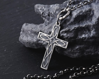 May include: A silver cross pendant necklace with an ornate, open design. The cross has a central emblem and curved details. The necklace chain is silver, with a textured, linked design. The background is a dark, textured surface, with a rough, stone-like appearance.